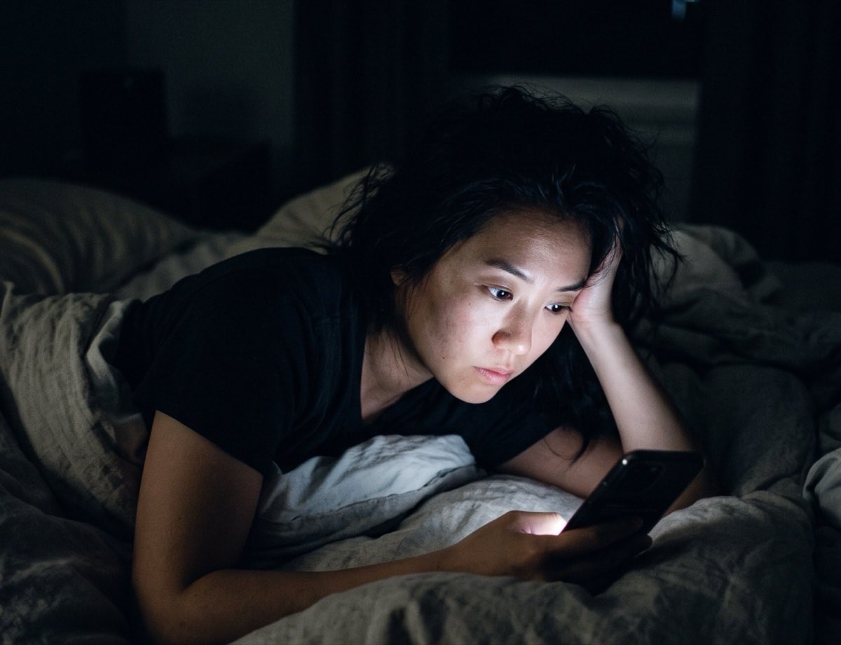 A woman lying in a dark bedroom, lit only by the phone she is reading.